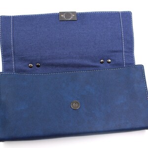 May include: Open, navy blue clutch with a denim-like interior. The clutch features a magnetic snap closure and a metal clasp. White stitching outlines the edges of the clutch.