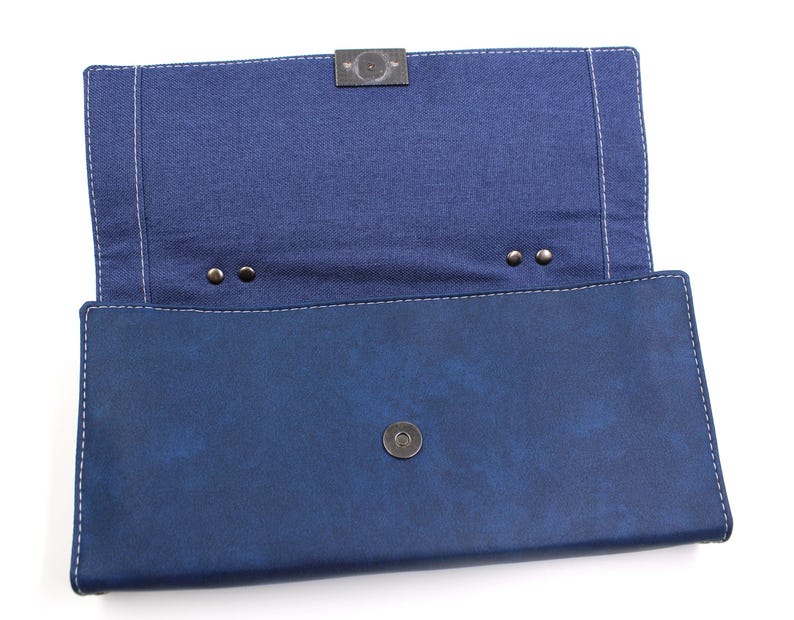 May include: Open, navy blue clutch with a denim-like interior. The clutch features a magnetic snap closure and a metal clasp. White stitching outlines the edges of the clutch.