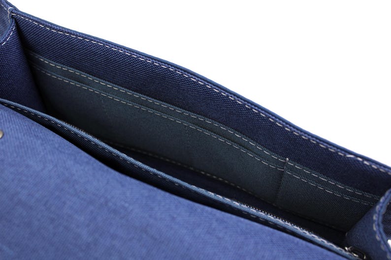 May include: Close-up of a blue denim handbag interior. The bag features multiple compartments with white stitching details. The interior is a darker shade of blue, and a zipper is visible at the bottom.