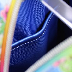 Puede incluir: Primer plano del interior de una peque&ntilde;a bolsa colorida. La bolsa tiene un forro de tela azul con un peque&ntilde;o bolsillo. El forro es visible a trav&eacute;s de la cremallera abierta.