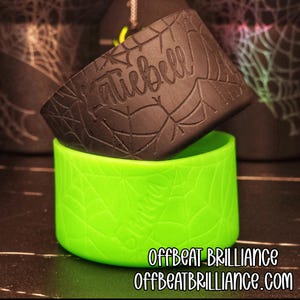 May include: A black water bottle sleeve with a spiderweb design and the name "Katiebell" is placed on top of a bright green water bottle sleeve. The text "Offbeat Brilliance" and the website address are at the bottom.