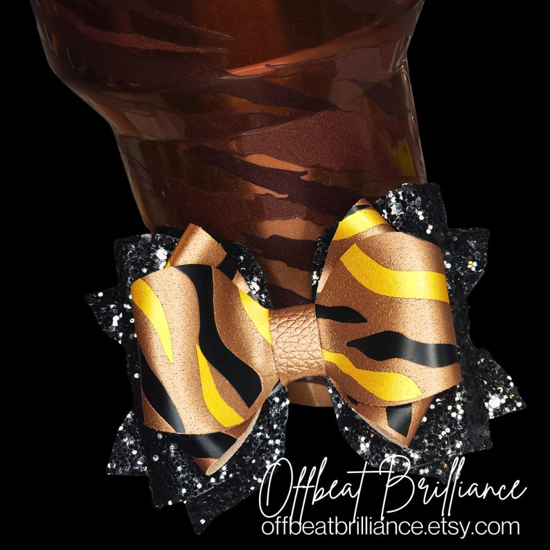 May include: A black and gold zebra print bow with black glitter trim. The bow is tied with a gold leather center.