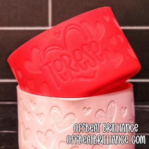 Könnte beinhalten: Eine rote Silikonhülle mit dem eingeprägten Namen "Terese", die auf einer rosa Keramiktasse liegt. Die Tasse hat ein Herzmuster und den Text "OFFBEAT BRILLIANCE" und "OFFBEATBRILLIANCE.COM".