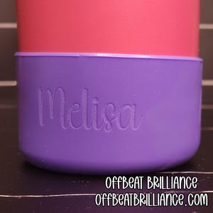 Może przedstawiać: Fioletowa osłona na kubek z wygrawerowanym imieniem "Melisa". Osłona jest owinięta wokół różowego kubka. Na dole obrazu znajduje się tekst "OFFBEAT BRILLIANCE" i "OFFBEATBRILLIANCE.com".