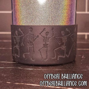 Może przedstawiać: Czarny rękaw na kubek z wzorami tańczących szkieletów. Sam kubek ma błyszczące, tęczowe wykończenie. Tekst "OFFBEAT BRILLIANCE" i "OFFBEATBRILLIANCE.COM" jest wydrukowany na dole.