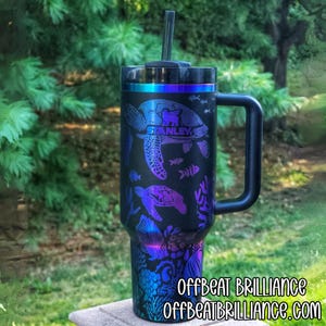 May include: Black Stanley tumbler with a handle, straw, and a sea turtle design. The design features sea turtles, fish, and coral in shades of blue, purple, and teal. The text "STANLEY" is visible on the tumbler. The bottom of the tumbler has the text "OFFBEAT BRILLIANCE" and "OFFBEATBRILLIANCE.COM".