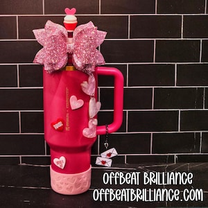 Könnte beinhalten: Ein rosa isolierter Becher mit Griff, verziert mit einer glitzernden rosa Schleife, herzförmigen Charms und herzförmigen Aufklebern. Der Boden des Bechers hat ein Herzmuster. Der Text "Offbeat Brilliance" und "Offbeatbrilliance.com" sind sichtbar.