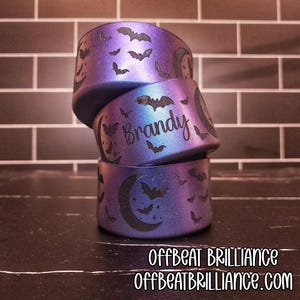 Peut inclure: Trois bracelets empilés, violets et bleus sur le thème d'Halloween, avec des motifs de chauves-souris et de lune. Le bracelet du milieu porte le nom "Brandy" gravé. Les bracelets portent le texte "OFFBEAT BRILLIANCE" et "OFFBEATBRILLIANCE.COM" en bas.