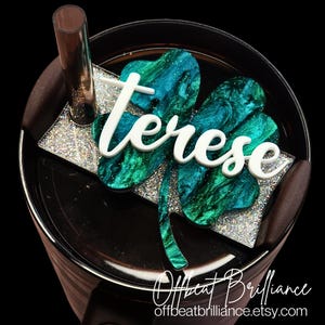 May include: A personalized tumbler topper featuring a four-leaf clover design in shades of teal and green with glitter accents. The name "Terese" is displayed in white script. The topper sits atop a black tumbler with a brown straw.