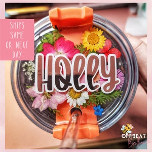 May include: A clear tumbler with a lid and straw, decorated with pressed flowers and the name "Holly" in gold lettering. The tumbler is filled with glitter and has a floral design.