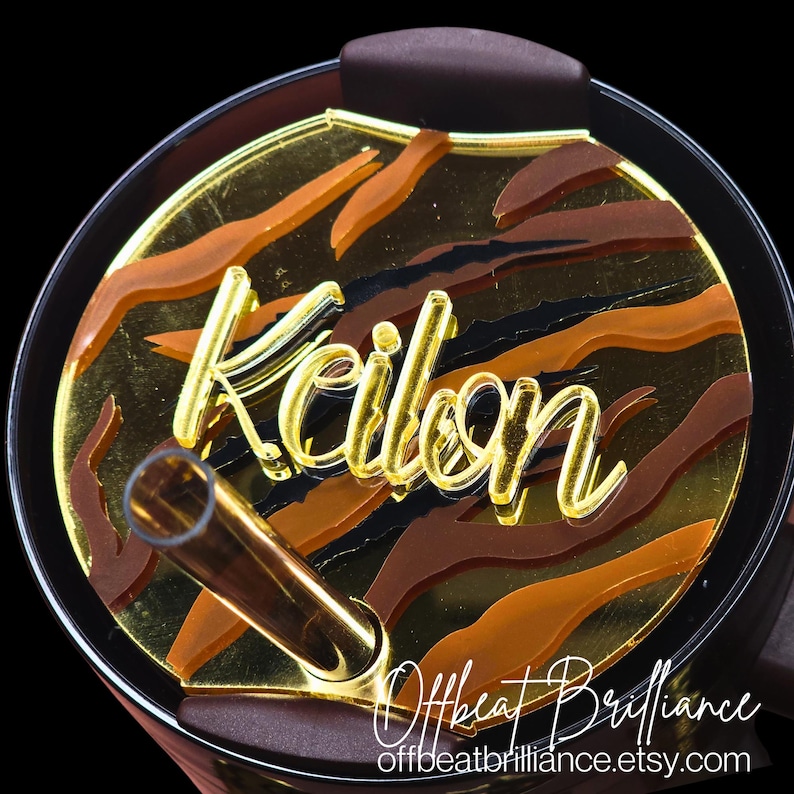 May include: A close-up of a black and gold tumbler lid with a tiger stripe pattern and the name "Kailyn" written in gold. A brown straw is sticking out of the lid.