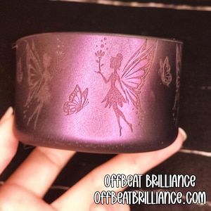 Pode incluir: Um recipiente cil&iacute;ndrico roxo com desenhos gravados de fadas e borboletas. O recipiente tem um brilho met&aacute;lico e apresenta o texto "OFFBEAT BRILLIANCE" e "OFFBEATBRILLIANCE.COM".