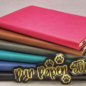 Puede incluir: Una pila de seis cuadernos con cubiertas de diferentes colores, incluyendo rosa, marr&oacute;n, gris, azul verdoso, azul y negro. Los cuadernos est&aacute;n apilados sobre una superficie negra con pegatinas doradas de huellas de patas y el texto "Pyr Paper Studio".