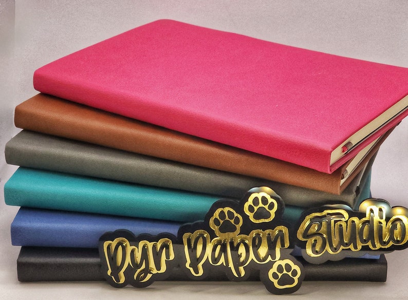 Puede incluir: Una pila de seis cuadernos con cubiertas de diferentes colores, incluyendo rosa, marr&oacute;n, gris, azul verdoso, azul y negro. Los cuadernos est&aacute;n apilados sobre una superficie negra con pegatinas doradas de huellas de patas y el texto "Pyr Paper Studio".
