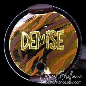 May include: A gold-colored, personalized tumbler lid with a tiger stripe pattern and the name "DEMISE" in gold letters. The lid has a black heart symbol and black claw marks.