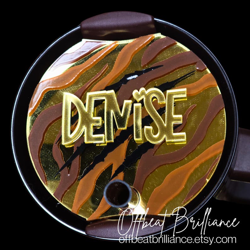 May include: A gold-colored, personalized tumbler lid with a tiger stripe pattern and the name "DEMISE" in gold letters. The lid has a black heart symbol and black claw marks.