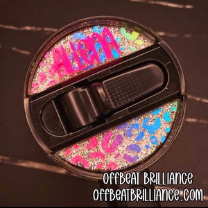 Puede incluir: Tapa de vaso negra con un diseño de estampado de leopardo brillante en rosa, azul y verde. El nombre "ALOHA" está en letras rosas y azules. El texto "OFFBEAT BRILLIANCE" y "OFFBEATBRILLIANCE.com" son en blanco.