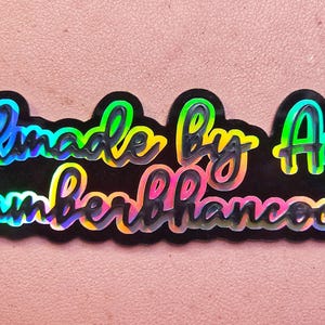 Puede incluir: Una pegatina negra con letras arco&iacute;ris iridiscentes que dice "Handmade by Amber @AmberHammock".