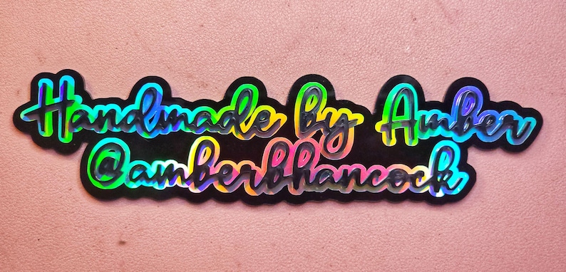Puede incluir: Una pegatina negra con letras arco&iacute;ris iridiscentes que dice "Handmade by Amber @AmberHammock".