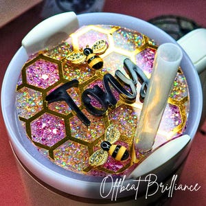 Könnte beinhalten: Ein weißer Becher mit einem rosa-goldenen Glitzerdeckel mit einem Wabenmuster, zwei Bienenanhängern und dem Namen "Tony" in schwarzen Buchstaben. Der Deckel hat auch einen durchsichtigen Plastikstrohhalm.