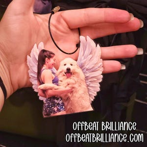 Pode incluir: Um pingente personalizado com uma mulher e um grande cão branco com asas de anjo. O pingente é suspenso por um cordão preto. O texto "Offbeat Brilliance" e "OffbeatBrilliance.com" são impressos na parte inferior.