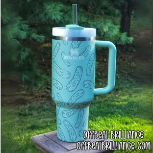 Puede incluir: Un vaso Stanley turquesa con asa y pajita, con un estampado de ilustraciones de pepinillos negros. El vaso tiene una tapa blanca y el texto "STANLEY" es visible. La esquina inferior derecha de la imagen tiene el texto "OFFBEAT BRILLIANCE" y "OFFBEATBRILLIANCE.COM".
