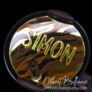 May include: A gold and brown patterned round lid with the name "SIMON" in gold letters. The lid is for a tumbler or cup.