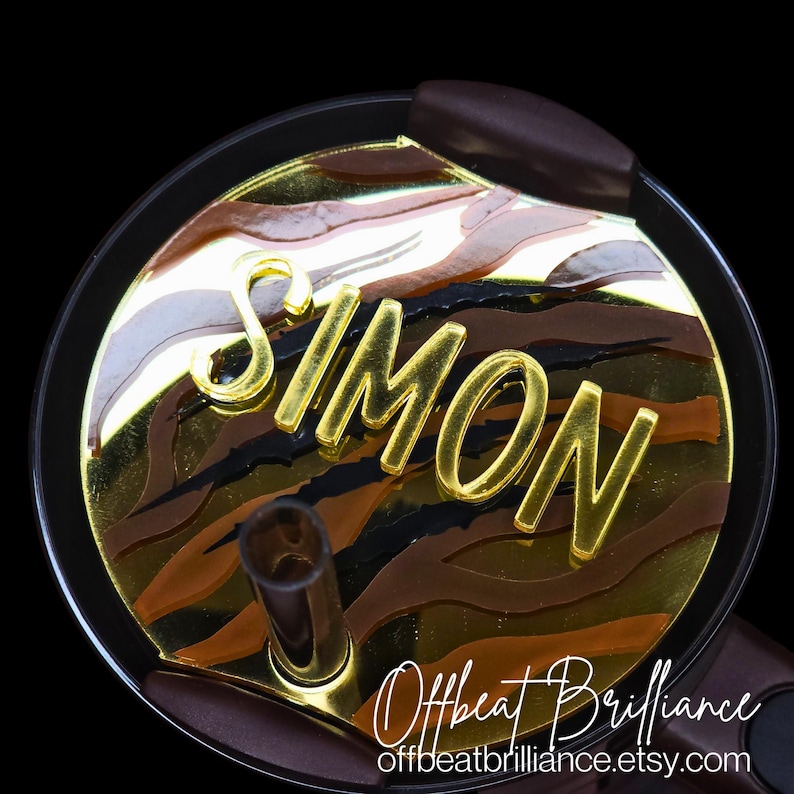 May include: A gold and brown patterned round lid with the name "SIMON" in gold letters. The lid is for a tumbler or cup.