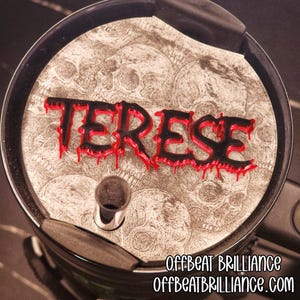 May include: A black and red lid with the name "TERESE" in a dripping font. The lid has a background of skulls and is part of a container. The website "OFFBEAT BRILLIANCE" is printed on the bottom right.