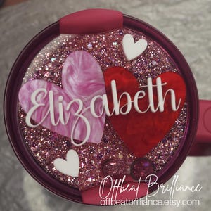 Könnte beinhalten: Ein rosa und roter Glitzerbecher mit einem weißen Herzen und dem Namen "Elizabeth" in weiß auf der Vorderseite.