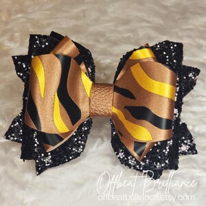 May include: A large black and gold bow with a zebra print pattern. The bow is made of fabric and has a glittery finish. The bow is tied with a brown leather strap. The bow is on a white background. Offbeat Brilliance offbeatbrilliance.etsy.com