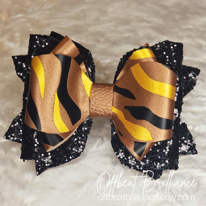 May include: A large black and gold bow with a zebra print pattern. The bow is made of fabric and has a glittery finish. The bow is tied with a brown leather strap. The bow is on a white background. Offbeat Brilliance offbeatbrilliance.etsy.com