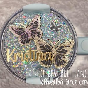 May include: A glittery tumbler lid with two butterfly designs and the name "Kristina" in gold lettering. The lid is light blue and has a handle. The text "OFFBEAT BRILLIANCE" and website address are visible.