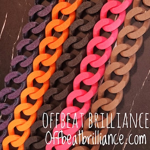 May include: Close-up of five colorful chunky chain necklaces in purple, orange, black, pink, and brown. The chains are arranged in parallel rows. The text "OFFBEAT BRILLIANCE Offbeatbrilliance.com" is visible in the lower right corner.