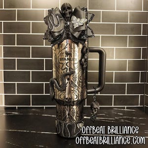 May include: A stainless steel Stanley tumbler with black embellishments. The tumbler features a black handle and is decorated with a black snake, skulls, and a bow with a butterfly. The text "STANLEY" is visible on the tumbler.