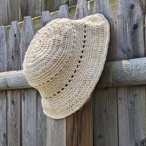 May include: A beige crocheted sun hat with a wide brim, hanging on a wooden fence.