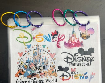 Op de afbeelding: Een doorzichtige plastic map met kleurrijke metalen ringen. De map bevat verschillende kleurrijke Disney-stickers, waaronder een kasteel, Mickey Mouse en de woorden "Disney Here We Come".