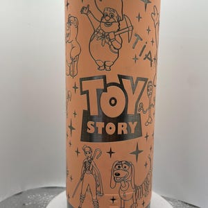May include: A stainless steel tumbler with a peach-colored exterior featuring black line art of characters from the movie "Toy Story." The words "Toy Story" are prominently displayed. The tumbler has a silver top and bottom.