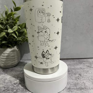 Puede incluir: Un vaso de acero inoxidable color crema con personajes de dibujos animados grabados y detalles de estrellas. El vaso tiene una tapa y base plateadas. El dise&ntilde;o incluye varios personajes de dibujos animados. El vaso se muestra sobre una plataforma blanca.