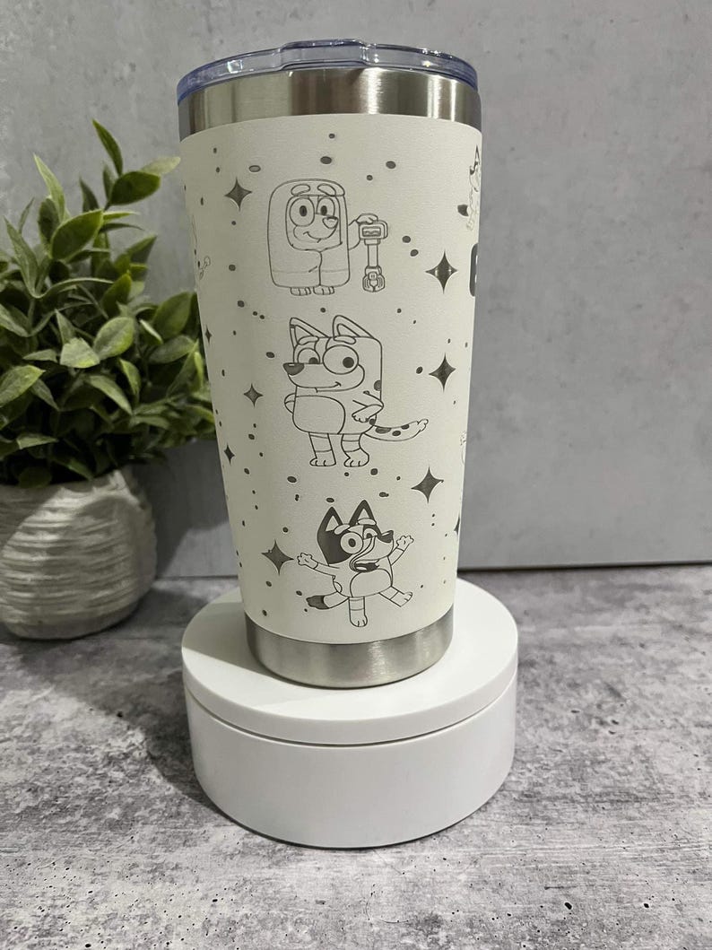 Puede incluir: Un vaso de acero inoxidable color crema con personajes de dibujos animados grabados y detalles de estrellas. El vaso tiene una tapa y base plateadas. El dise&ntilde;o incluye varios personajes de dibujos animados. El vaso se muestra sobre una plataforma blanca.