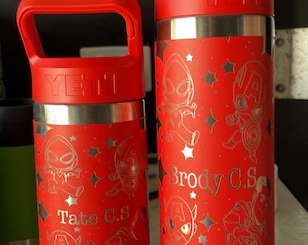 Puede incluir: Dos botellas de agua rojas de la marca Yeti con acabado de acero inoxidable. Las botellas tienen un asa y están decoradas con un diseño plateado de superhéroes de dibujos animados y estrellas. La botella más pequeña tiene el nombre "Tate C.S." grabado en ella, y la botella más grande tiene el nombre "Brody C.S." grabado en ella.