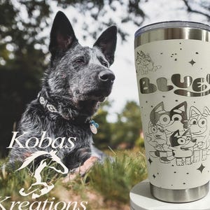 May include: A stainless steel tumbler with a cream-colored finish, featuring a Bluey cartoon design and the word "Bluey". The tumbler is decorated with stars and cartoon characters. A dog is in the background.