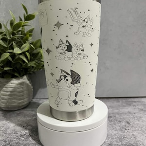 Puede incluir: Un vaso aislado blanco con personajes de dibujos animados de perros y estrellas. El vaso de acero inoxidable tiene una tapa y una base plateadas. El dise&ntilde;o est&aacute; grabado en la superficie del vaso. Dise&ntilde;ado para mantener las bebidas fr&iacute;as o calientes.
