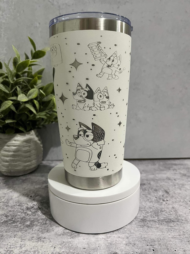 Puede incluir: Un vaso aislado blanco con personajes de dibujos animados de perros y estrellas. El vaso de acero inoxidable tiene una tapa y una base plateadas. El dise&ntilde;o est&aacute; grabado en la superficie del vaso. Dise&ntilde;ado para mantener las bebidas fr&iacute;as o calientes.