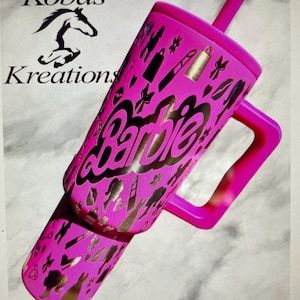 Puede incluir: Un vaso rosa con asa y pajita, con la palabra "Barbie" en letras doradas. El vaso está decorado con siluetas doradas de varios objetos. El logotipo "Kobas Kreations" es visible en el fondo.