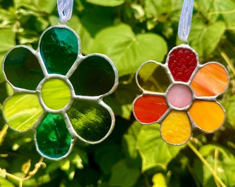 May include: Two stained glass flower ornaments with a silver metal outline and white ribbon for hanging. One flower is green and yellow, the other is red, orange, pink, and yellow. The background is green foliage.