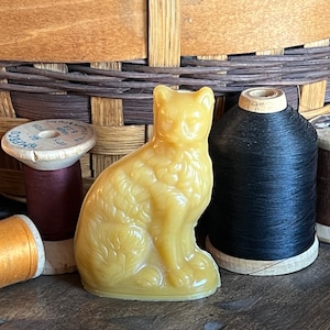 May include: A yellow cat figurine sits among spools of thread in various colors. The cat is detailed with textured fur. The spools include black, brown, purple, and gold thread. A woven basket is in the background.