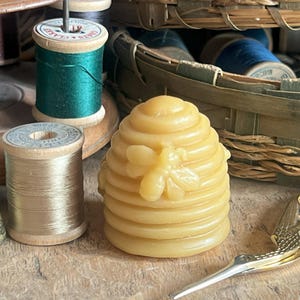 May include: A yellow beeswax candle shaped like a beehive with a bee on the front. The candle is surrounded by sewing supplies, including spools of thread in various colors, a thimble, and gold scissors.