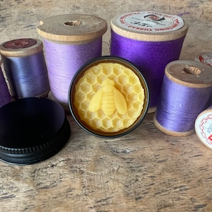 May include: A collection of purple thread spools surrounds a small, round container with a bee-shaped design. The container has a black lid. The spools are various shades of purple and lavender, with wooden sides.