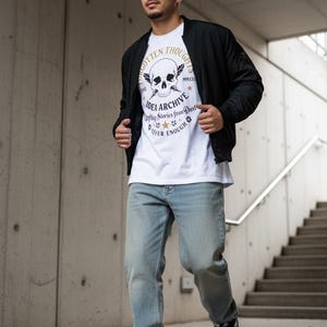 May include: A white t-shirt featuring a black graphic of a skull with wings and the text Forgotten Thoughts Idea Archive Stories from Dust Never Enough MMXXI, worn under a black bomber jacket with light blue denim jeans and black and white sneakers.